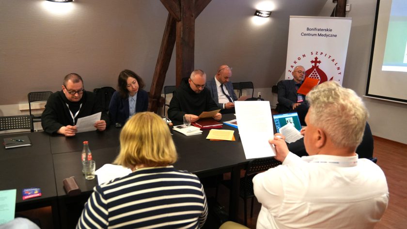 Uczestnicy spotkania siedzą przy dużym, ciemnym stole konferencyjnym w sali na poddaszu z drewnianymi belkami stropowymi. Kilka osób czyta wydrukowane materiały, a w tle widoczny jest baner Bonifraterskiego Centrum Medycznego z logo Zakonu Szpitalnego. Na pierwszym planie mężczyzna w białej koszuli trzyma dokument, obok siedzą inni uczestnicy spotkania skupieni na dyskusji i lekturze materiałów