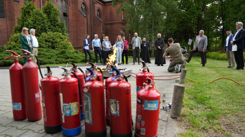 Na dziedzińcu przed ceglastym budynkiem uczestnicy szkolenia przeciwpożarowego obserwują pokaz gaszenia ognia. Na pierwszym planie ustawiono kilka czerwonych gaśnic, a pośrodku alejki płonie kontrolowane ognisko w metalowej kuwecie. Instruktor klęczy obok źródła ognia i prezentuje zasady użycia sprzętu gaśniczego, podczas gdy grupa uczestników, w tym osoby świeckie i duchowne, stoi w półkolu i przygląda się demonstracji