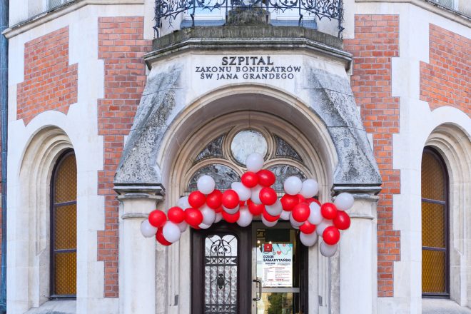 Wejście do Szpitala Zakonu Bonifratrów św. Jana Grandego w Krakowie przy ul. Trynitarskiej 11. Nad drzwiami widnieje napis z nazwą szpitala, a wejście udekorowane jest girlandą z czerwonych i białych balonów. Na szybie drzwi znajduje się plakat zapowiadający Dzień Samarytański organizowany przez Bonifraterskie Centrum Medyczne