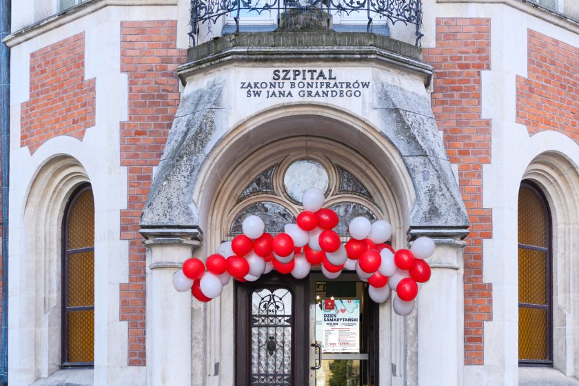 Wejście do Szpitala Zakonu Bonifratrów św. Jana Grandego w Krakowie przy ul. Trynitarskiej 11. Nad drzwiami widnieje napis z nazwą szpitala, a wejście udekorowane jest girlandą z czerwonych i białych balonów. Na szybie drzwi znajduje się plakat zapowiadający Dzień Samarytański organizowany przez Bonifraterskie Centrum Medyczne