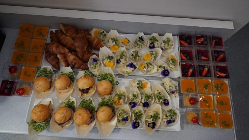 Bufet z przekąskami przygotowany dla uczestników wydarzenia. Na stole znajdują się mini burgery, croissanty, sałatki i desery w pucharkach