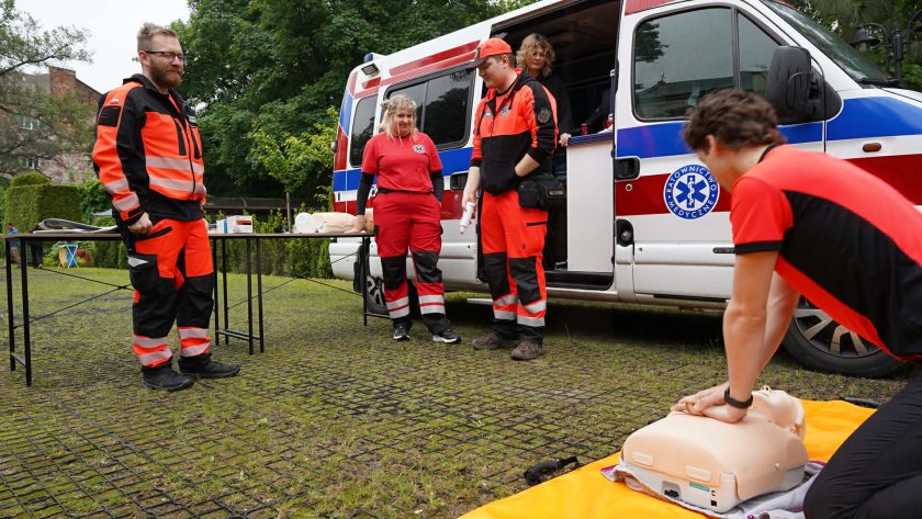 Ratownicy medyczni prowadzą pokaz resuscytacji krążeniowo-oddechowej z wykorzystaniem fantoma. Obok stoi ambulans oraz uczestnicy szkolenia