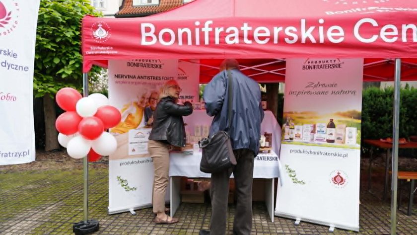 Stoisko Produktów Bonifraterskich pod czerwonym namiotem Bonifraterskiego Centrum Medycznego. Pracownica rozmawia z odwiedzającym przy ekspozycji naturalnych preparatów zdrowotnych