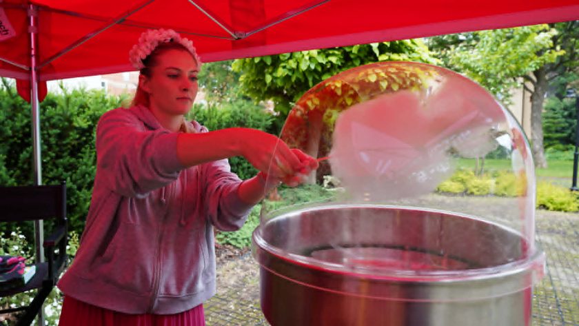 Młoda wolontariuszka przygotowuje watę cukrową przy specjalnej maszynie ustawionej pod czerwonym namiotem podczas plenerowego wydarzenia organizowanego przez Bonifraterskie Centrum Medyczne