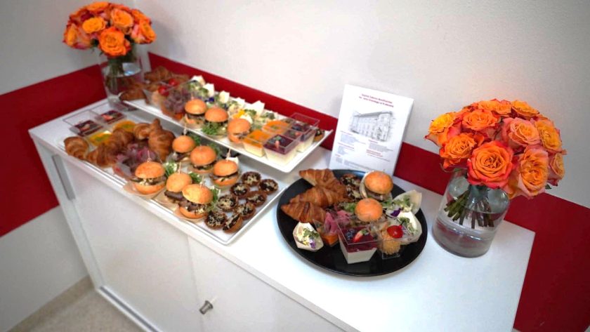 Stół cateringowy przygotowany dla uczestników wydarzenia. Na półmiskach znajdują się mini burgery, croissanty, przekąski, sałatki i desery, a całość uzupełniają bukiety pomarańczowych róż