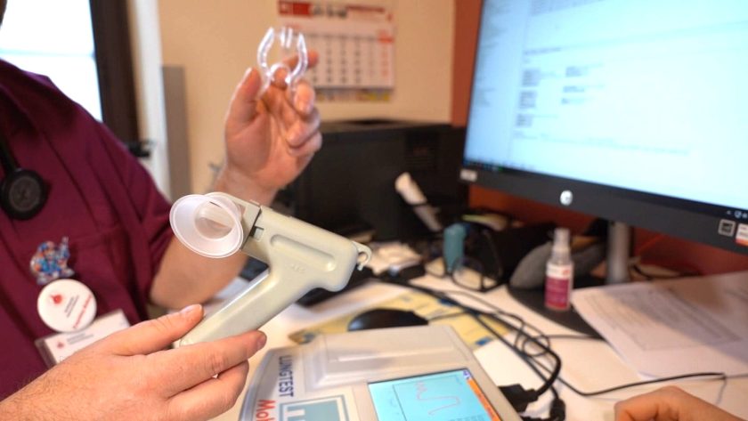 Pracownik medyczny prezentuje pacjentce ustnik wykorzystywany podczas badania spirometrycznego. Na biurku obok znajduje się spirometr oraz monitor z wynikami pomiarów
