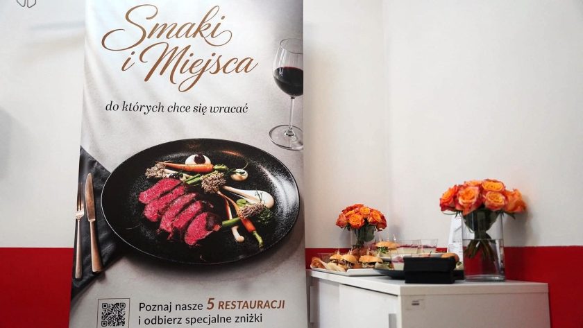 Roll-up promujący akcję „Smaki i Miejsca, do których chce się wracać” ustawiony przy stoliku z poczęstunkiem. Obok znajdują się kompozycje z pomarańczowych róż oraz przekąski przygotowane dla uczestników wydarzenia