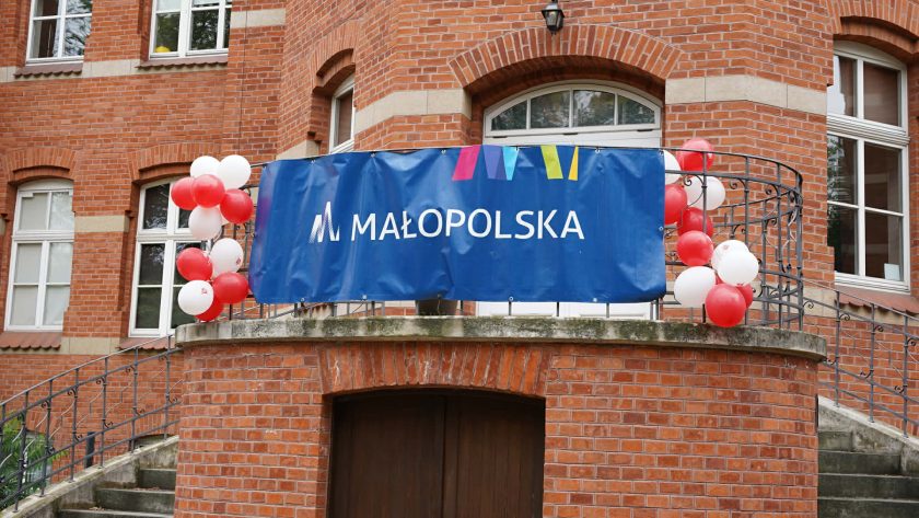 Baner Województwa Małopolskiego zawieszony na balkonie zabytkowego ceglanego budynku szpitalnego. Balustradę udekorowano biało-czerwonymi balonami nawiązującymi do wydarzenia organizowanego przez Bonifraterskie Centrum Medyczne