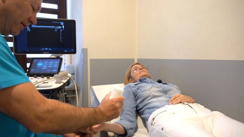 Lekarz wykonuje badanie ultrasonograficzne dłoni u pacjentki leżącej na kozetce w gabinecie diagnostycznym. Obok znajduje się aparat USG z monitorem wyświetlającym obraz badania