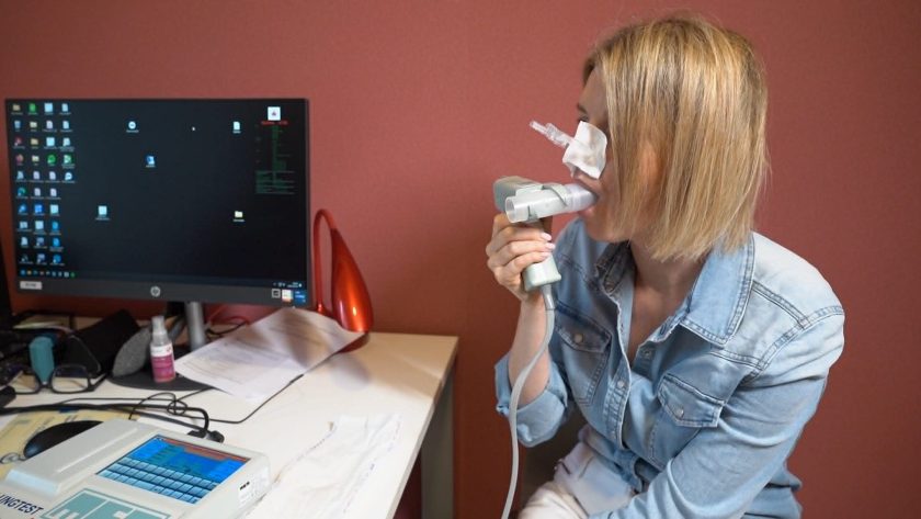 Pacjentka siedzi przy stanowisku diagnostycznym podczas badania spirometrycznego. Trzyma ustnik spirometru w ustach, a na nosie ma założony specjalny klips umożliwiający pomiar wydolności oddechowej