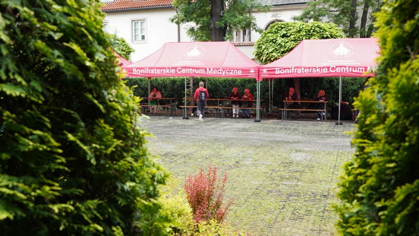 Widok na dziedziniec z czerwonymi namiotami Bonifraterskiego Centrum Medycznego. Pod zadaszeniem przy stołach siedzą uczestnicy wydarzenia, a całość otaczają zadbane krzewy i drzewa