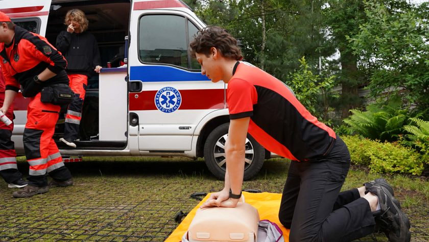 Ratownik medyczny wykonuje uciski klatki piersiowej na fantomie podczas pokazu pierwszej pomocy. W tle widoczny jest ambulans oraz pozostali członkowie zespołu ratownictwa medycznego przygotowujący sprzęt do szkolenia