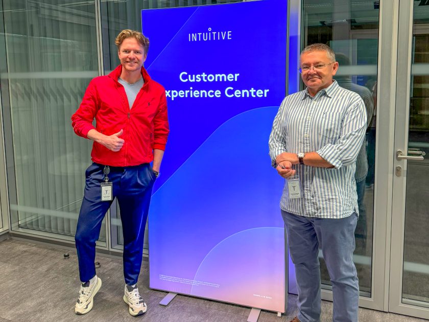 Dwóch mężczyzn stoi po obu stronach podświetlanego ekranu z napisem „INTUITIVE Customer Experience Center”. Po lewej mężczyzna w czerwonej kurtce wykonuje gest uniesionego kciuka, po prawej mężczyzna w biało-niebieskiej koszuli w paski. Zdjęcie wykonano we wnętrzu nowoczesnego centrum szkoleniowego z przeszklonymi ścianami