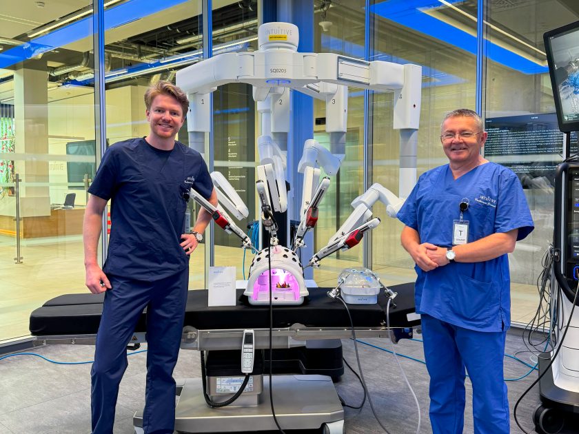 Dwóch mężczyzn w strojach medycznych stoi po obu stronach robota chirurgicznego da Vinci. Urządzenie z rozłożonymi ramionami operacyjnymi znajduje się na środku kadru. Po lewej instruktor w granatowym uniformie, po prawej uczestnik szkolenia w niebieskim stroju medycznym. Zdjęcie wykonano w centrum szkoleniowym firmy Intuitive podczas prezentacji technologii chirurgii robotycznej