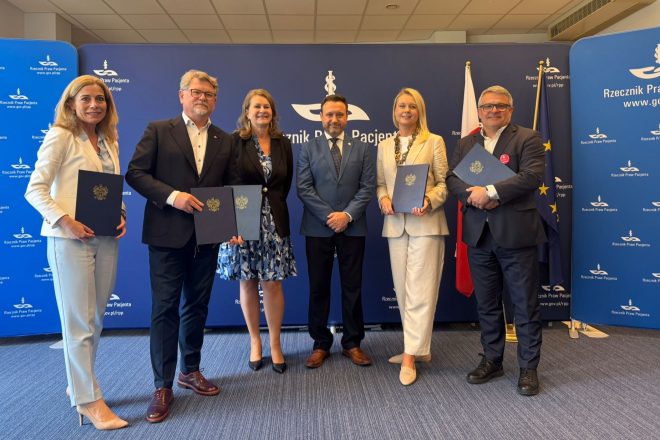 Sześcioro uczestników uroczystości pozuje do wspólnego zdjęcia przed granatową ścianką Rzecznika Praw Pacjenta. Cztery osoby trzymają granatowe teczki z godłem państwowym. W centrum stoi mężczyzna w niebieskim garniturze, po jego bokach ustawione są kobiety i mężczyźni w strojach biznesowych. Po prawej stronie widoczne są flagi Polski i Unii Europejskiej. Zdjęcie wykonano w sali konferencyjnej podczas oficjalnego wydarzenia