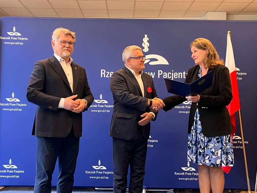 Kobieta w czarnej marynarce i wzorzystej sukience wręcza dokument oraz ściska dłoń mężczyzny w ciemnym garniturze. Obok stoi drugi mężczyzna w granatowym garniturze, obserwujący ceremonię. W tle znajduje się granatowa ścianka z logo Rzecznika Praw Pacjenta oraz flaga Polski. Zdjęcie przedstawia moment oficjalnego przekazania dokumentu podczas uroczystości