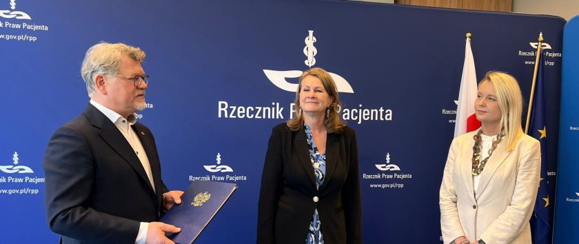Przed granatową ścianką z logotypem Rzecznika Praw Pacjenta stoi troje uczestników uroczystości. Mężczyzna w ciemnym garniturze i jasnej koszuli przemawia, trzymając w dłoniach granatową teczkę z godłem państwowym. Obok niego stoją dwie kobiety w formalnych strojach, słuchające wystąpienia. W tle widoczne są flagi Polski i Unii Europejskiej oraz duże logo Rzecznika Praw Pacjenta