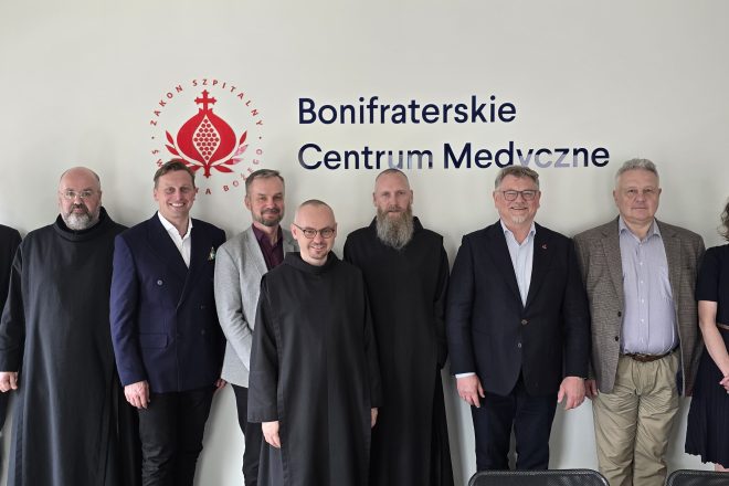 Zdjęcie obrazuje grupę dziewięciu osób pozujących do wspólnej fotografii na tle jasnej ściany z dużym logo i napisem „Bonifraterskie Centrum Medyczne” oraz czerwonym symbolem Zakonu Szpitalnego św. Jana Bożego. Wśród uczestników znajdują się zarówno osoby świeckie ubrane w garnitury i stroje biznesowe, jak i trzej zakonnicy bonifratrzy w charakterystycznych czarnych habitach. W pierwszym rzędzie, na środku, stoi młody zakonnik w okularach, natomiast pozostali uczestnicy ustawieni są w jednej linii za nim. Większość osób uśmiecha się do obiektywu, tworząc oficjalny, lecz przyjazny charakter zdjęcia. W dolnej części kadru widoczne są fragmenty oparć krzeseł, a wnętrze jest nowoczesne, dobrze oświetlone światłem dziennym i utrzymane w jasnej kolorystyce
