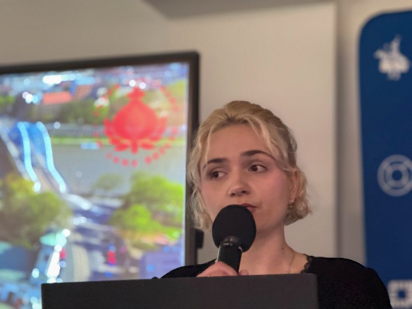 Zbliżenie na prelegentkę występującą podczas konferencji. Kobieta trzyma mikrofon i przemawia do uczestników wydarzenia, a w tle widoczny jest ekran z prezentacją oraz logo Szpitala Bonifratrów w Krakowie