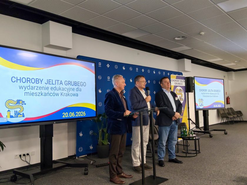 Otwarcie wydarzenia edukacyjnego „Choroby jelita grubego” dla mieszkańców Krakowa. Trzech mężczyzn stoi na scenie przed ekranem prezentacyjnym, witając uczestników konferencji