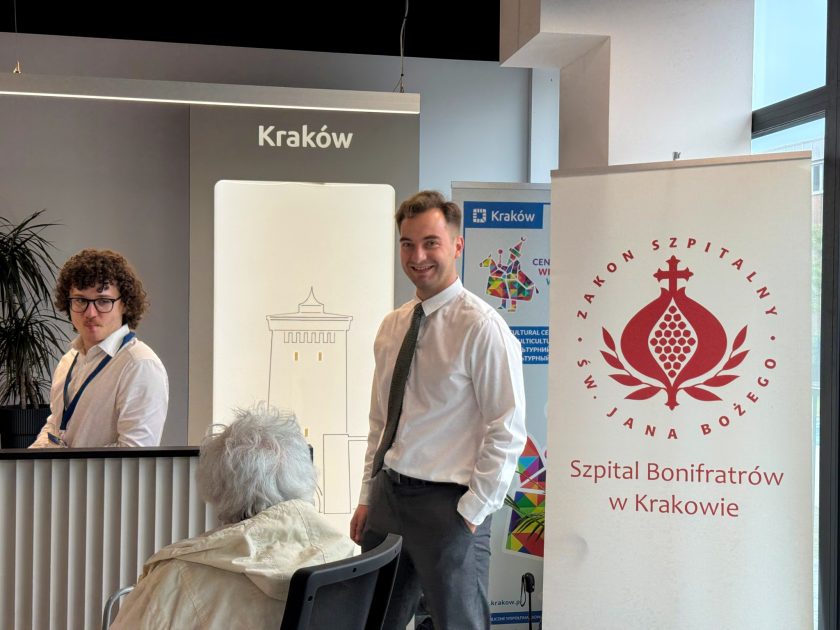 Uczestnicy i organizatorzy wydarzenia rozmawiają w przestrzeni konferencyjnej. Obok stanowiska informacyjnego widoczny jest roll-up Szpitala Bonifratrów w Krakowie
