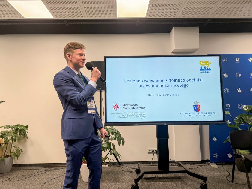Dr n. med. Paweł Bogacki prowadzi wykład dotyczący utajonego krwawienia z dolnego odcinka przewodu pokarmowego. Prelegent stoi przy ekranie prezentacyjnym i omawia zagadnienia związane z profilaktyką oraz diagnostyką chorób jelita grubego
