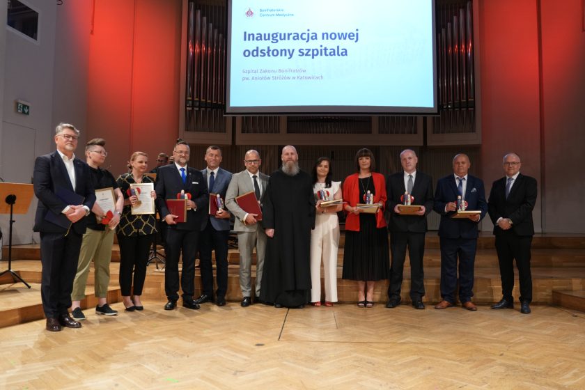 Zdjęcie przedstawia grupę laureatów oraz organizatorów stojących na scenie sali koncertowej podczas uroczystości inauguracji nowej odsłony Szpitala Zakonu Bonifratrów pw. Aniołów Stróżów w Katowicach. Każda z wyróżnionych osób trzyma pamiątkową statuetkę lub dyplom. W centrum stoi zakonnik Zakonu Bonifratrów w czarnym habicie, a po obu stronach znajdują się przedstawiciele instytucji i partnerów zaangażowanych w modernizację szpitala. Nad uczestnikami widoczny jest ekran z napisem „Inauguracja nowej odsłony szpitala”, a w tle znajdują się organy koncertowe oraz muzycy orkiestry