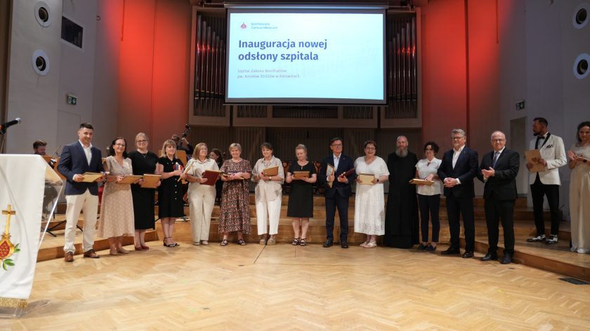 Zdjęcie przedstawia grupę pracowników szpitala oraz organizatorów stojących na scenie podczas uroczystości inauguracji nowej odsłony Szpitala Zakonu Bonifratrów pw. Aniołów Stróżów w Katowicach. Wyróżnione osoby trzymają pamiątkowe dyplomy i statuetki, ustawione są w jednym rzędzie. Po prawej stronie stoją przedstawiciele władz Bonifraterskiego Centrum Medycznego oraz zakonnik Zakonu Bonifratrów. W tle widoczny jest ekran z nazwą wydarzenia, organy koncertowe oraz członkowie zespołu muzycznego przygotowującego oprawę artystyczną