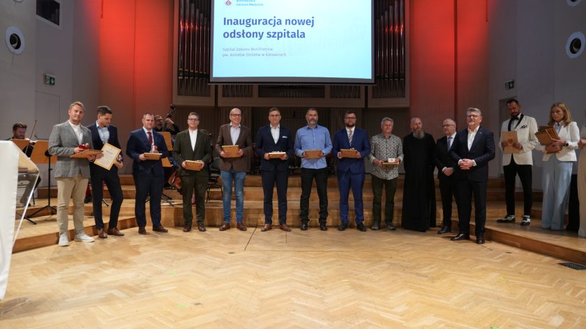 Zdjęcie przedstawia grupę przedstawicieli firm i instytucji wyróżnionych podczas inauguracji nowej odsłony Szpitala Zakonu Bonifratrów pw. Aniołów Stróżów w Katowicach. Uczestnicy stoją na scenie, trzymając pamiątkowe statuetki i dyplomy. Obok nich stoją przedstawiciele Bonifraterskiego Centrum Medycznego oraz zakonnik Zakonu Bonifratrów. W tle znajduje się ekran z nazwą wydarzenia oraz organy koncertowe, a po bokach sceny widoczni są prowadzący uroczystość