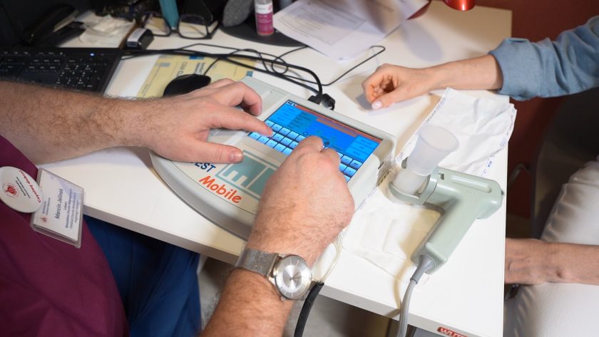 Zbliżenie na dłonie lekarza obsługującego ekran dotykowy spirometru. Obok urządzenia leży ustnik przygotowany do badania oddechowego