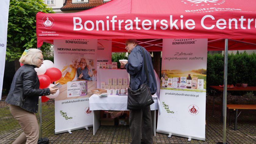 Stoisko Produktów Bonifraterskich pod czerwonym namiotem podczas wydarzenia plenerowego. Odwiedzający zapoznają się z ofertą suplementów, kosmetyków i preparatów ziołowych