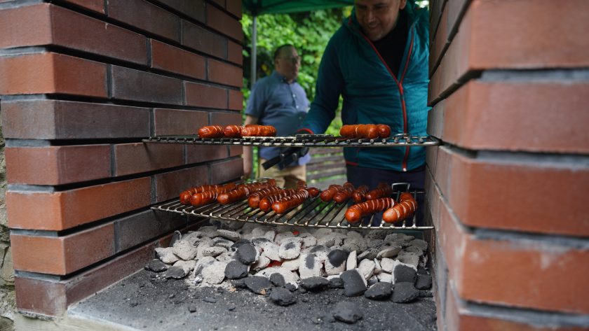 Kiełbaski pieką się na ruszcie nad rozżarzonym węglem w murowanym grillu. Mężczyzna dogląda przygotowywanego poczęstunku