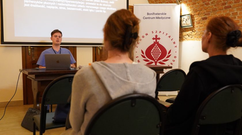 Młody prelegent prowadzi wykład w sali konferencyjnej Bonifraterskiego Centrum Medycznego. Uczestnicy słuchają prezentacji dotyczącej prawidłowego oddychania