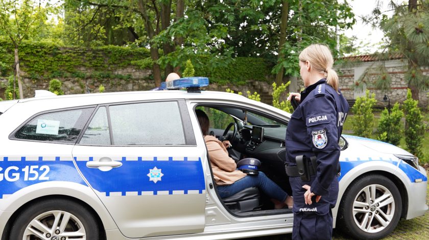 Policjantka stoi obok oznakowanego radiowozu policyjnego z otwartymi drzwiami kierowcy. Kobieta siedząca za kierownicą zapoznaje się z wyposażeniem pojazdu podczas wydarzenia plenerowego
