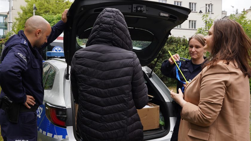 Policjanci prezentują wyposażenie radiowozu uczestnikom wydarzenia plenerowego. Przy otwartym bagażniku funkcjonariuszka pokazuje elementy wyposażenia służbowego