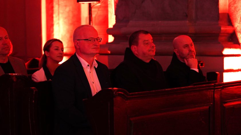 Publiczność siedzi w ławkach kościoła i słucha koncertu. Wnętrze świątyni oświetlone jest czerwonym światłem scenicznym