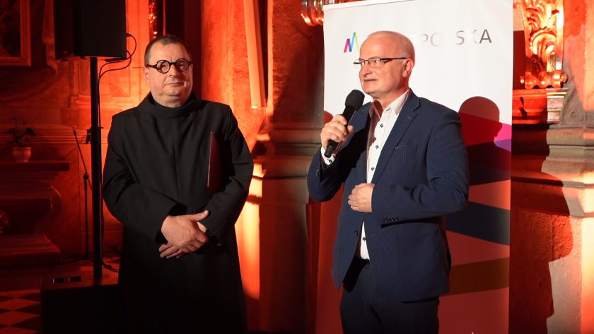 Dwóch mężczyzn przemawia do zgromadzonych uczestników wydarzenia. Jeden z nich trzyma mikrofon, drugi stoi obok w habicie zakonnym na tle baneru Małopolski