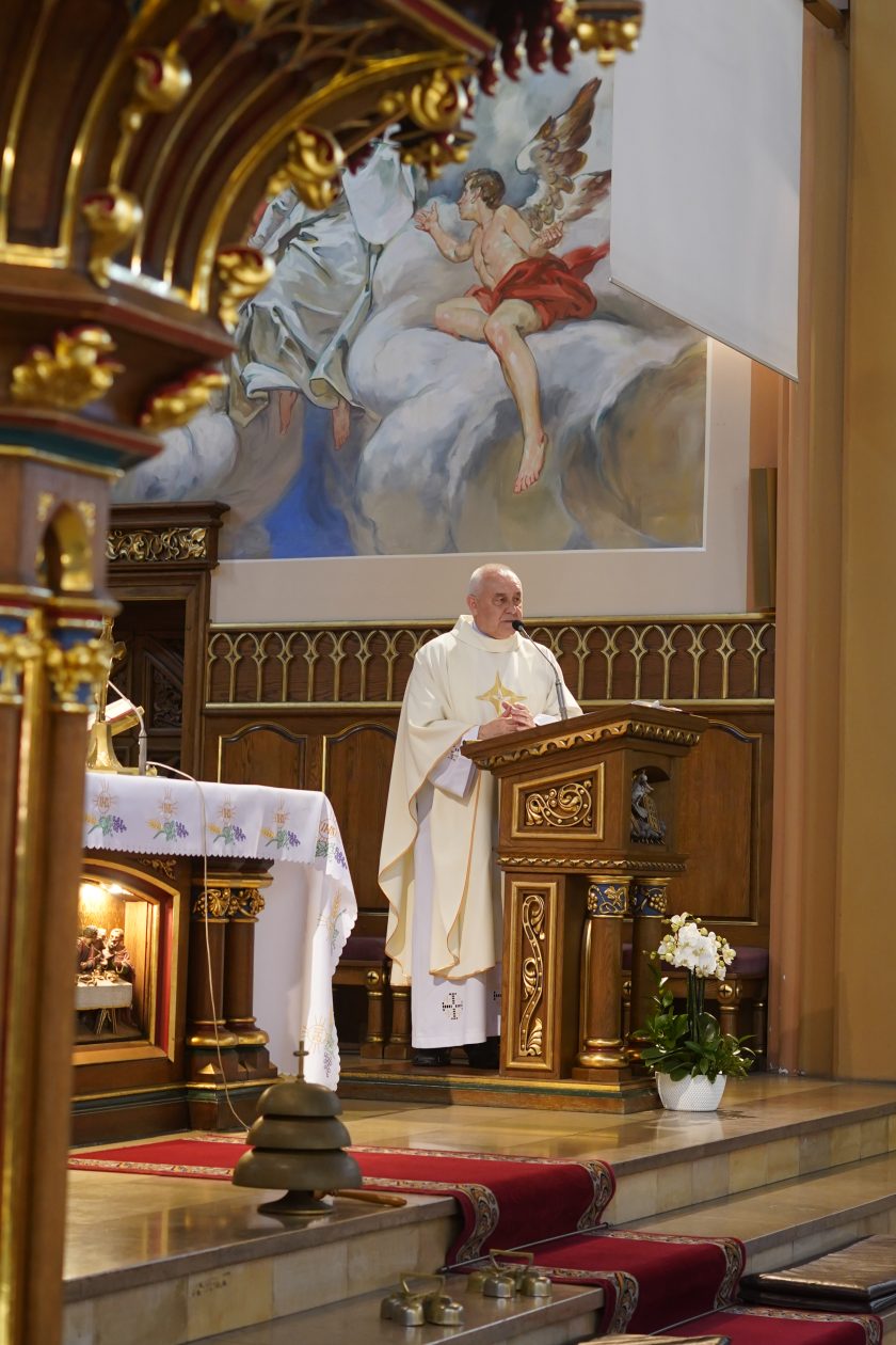 Zdjęcie przedstawia kapłana ubranego w białe szaty liturgiczne, który wygłasza homilię z bogato zdobionej drewnianej ambony podczas uroczystej mszy świętej w kościele Zakonu Bonifratrów. W tle znajduje się duży mural przedstawiający scenę religijną z aniołem oraz postaciami na obłokach. Obok ambony ustawiona jest biała doniczka z kwitnącym storczykiem, a po lewej stronie widoczny jest ołtarz przykryty haftowanym obrusem. Wnętrze świątyni utrzymane jest w ciepłych odcieniach drewna i złotych zdobień