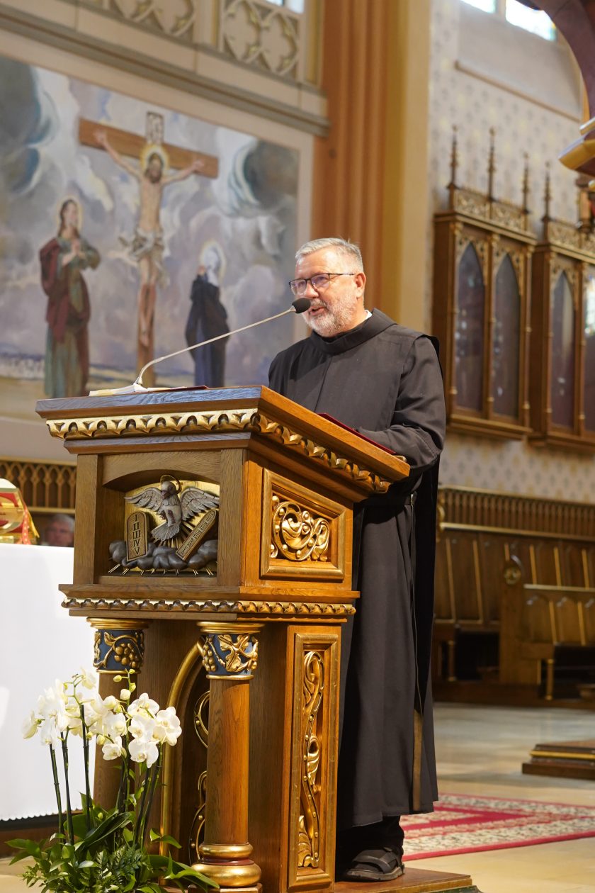 Zdjęcie przedstawia zakonnika Zakonu Bonifratrów ubranego w czarny habit, przemawiającego z drewnianej, bogato zdobionej ambony podczas uroczystej mszy świętej. Na froncie ambony znajduje się płaskorzeźba z symbolem Ducha Świętego. W tle widoczne są obrazy przedstawiające sceny Męki Pańskiej oraz drewniane elementy wyposażenia kościoła. Przed amboną ustawione są białe storczyki, a wnętrze świątyni rozświetla ciepłe światło wpadające przez okna