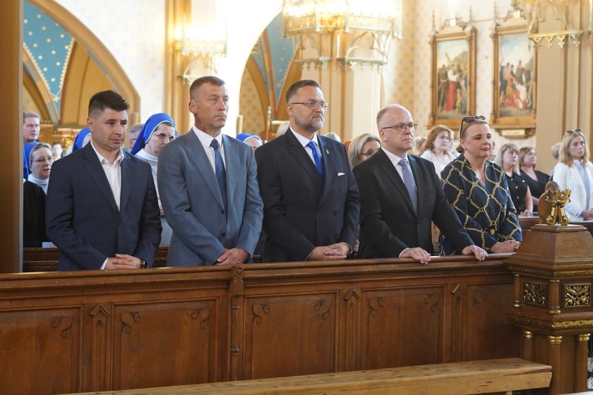 Zdjęcie przedstawia przedstawicieli władz samorządowych i instytucji stojących w pierwszej ławce podczas uroczystej mszy świętej. Mężczyźni ubrani są w garnitury, a kobieta w elegancki granatowy strój z ozdobnym złotym wzorem. Za nimi widoczne są siostry zakonne oraz pozostali uczestnicy nabożeństwa. W tle znajdują się gotyckie łuki świątyni, obrazy Drogi Krzyżowej oraz dekoracyjne oświetlenie kościoła