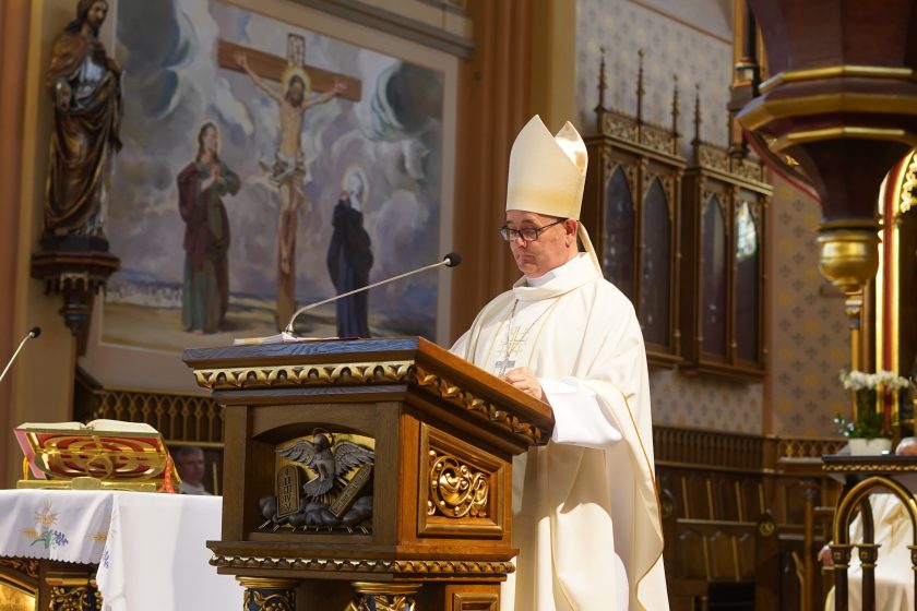 Zdjęcie przedstawia biskupa ubranego w białe szaty liturgiczne oraz mitrę, który odczytuje przygotowany tekst z drewnianej ambony podczas uroczystej mszy świętej. Na pierwszym planie widoczny jest fragment ołtarza z otwartą księgą liturgiczną, natomiast w tle znajduje się malowidło przedstawiające ukrzyżowanego Chrystusa oraz świętych. Wnętrze kościoła utrzymane jest w jasnych kolorach z licznymi drewnianymi elementami wyposażenia
