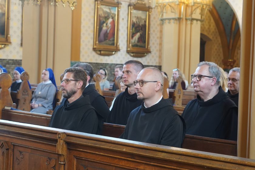 Zdjęcie przedstawia czterech zakonników Zakonu Bonifratrów siedzących w drewnianych ławkach podczas uroczystej mszy świętej. Bracia zakonni ubrani są w czarne habity i z uwagą uczestniczą w liturgii. W tle widoczne są siostry zakonne w niebiesko-szarych habitach oraz pozostali uczestnicy nabożeństwa. Ściany kościoła zdobią obrazy Drogi Krzyżowej oraz drewniane elementy wyposażenia świątyni