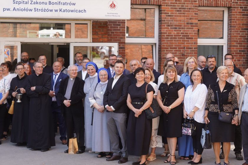 Zdjęcie przedstawia grupę uczestników uroczystości zgromadzonych przed wejściem do Szpitala Zakonu Bonifratrów pw. Aniołów Stróżów w Katowicach. W pierwszym rzędzie stoją przedstawiciele władz, siostry zakonne, pracownicy szpitala oraz zaproszeni goście. Po lewej stronie widoczny jest zakonnik trzymający kropidło, obok którego stoją duchowni oraz przedstawiciele Bonifraterskiego Centrum Medycznego. Nad wejściem znajduje się tablica z nazwą szpitala i logo Bonifraterskiego Centrum Medycznego. W tle widoczna jest ceglana elewacja budynku oraz pozostali uczestnicy wydarzenia ustawieni do wspólnego zdjęcia