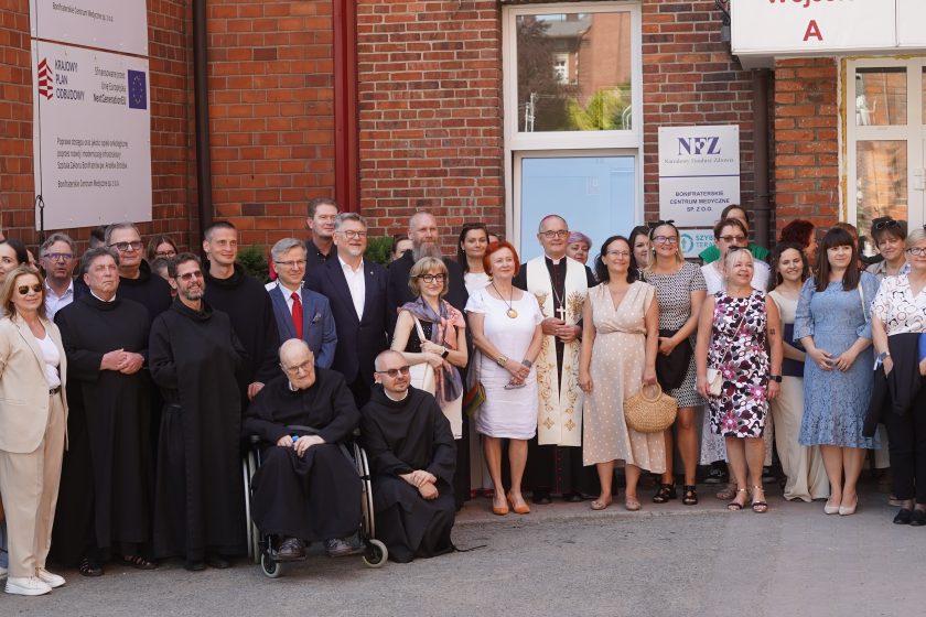 Zdjęcie przedstawia liczną grupę osób zgromadzonych przed głównym wejściem do Szpitala Zakonu Bonifratrów pw. Aniołów Stróżów w Katowicach. Na pierwszym planie stoją i siedzą zakonnicy Zakonu Bonifratrów, duchowni oraz przedstawiciele szpitala i zaproszeni goście. W centrum widoczny jest biskup ubrany w białe szaty liturgiczne z pastorałem, obok którego stoją uczestnicy uroczystości w eleganckich strojach. Przed grupą siedzi starszy zakonnik na wózku inwalidzkim oraz młodszy brat zakonny. W tle znajduje się ceglana elewacja szpitala z tablicami informacyjnymi, w tym tablica dotycząca dofinansowania inwestycji z Krajowego Planu Odbudowy oraz oznaczenia Narodowego Funduszu Zdrowia. Zdjęcie wykonano podczas wspólnej fotografii uczestników inauguracji po zakończeniu uroczystości