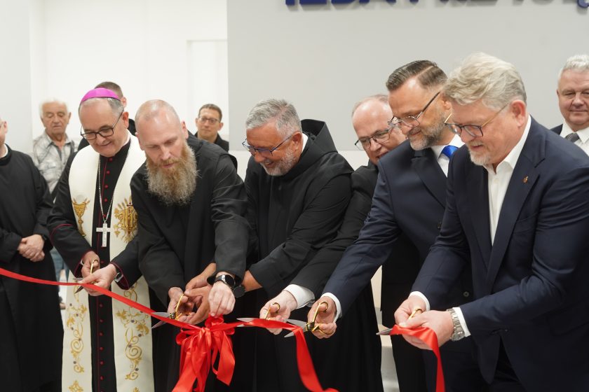 Zdjęcie przedstawia uroczyste przecięcie czerwonej wstęgi podczas otwarcia zmodernizowanej części Szpitala Zakonu Bonifratrów pw. Aniołów Stróżów w Katowicach. W wydarzeniu uczestniczą biskup w białych szatach liturgicznych z fioletową piuską, zakonnicy Zakonu Bonifratrów oraz przedstawiciele władz Bonifraterskiego Centrum Medycznego i zaproszeni goście w garniturach. Każda z osób trzyma złote nożyczki i jednocześnie przecina szeroką czerwoną wstęgę ozdobioną dużą kokardą. W tle widoczne są jasne ściany nowoczesnego wnętrza szpitala oraz pozostali uczestnicy wydarzenia obserwujący uroczystość