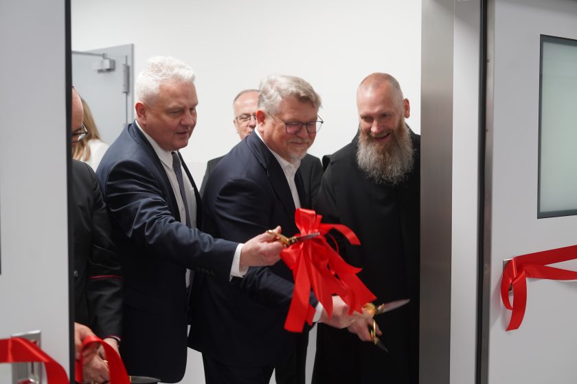 Zdjęcie przedstawia przedstawicieli Bonifraterskiego Centrum Medycznego oraz Zakonu Bonifratrów podczas symbolicznego otwarcia nowej części szpitala. Dwóch mężczyzn w garniturach i zakonnik w czarnym habicie jednocześnie przecinają czerwoną wstęgę rozpiętą w drzwiach prowadzących do nowo wyremontowanych pomieszczeń. Na środku znajduje się ozdobna czerwona kokarda. Wszyscy uczestnicy uśmiechają się, podkreślając uroczysty charakter wydarzenia. W tle widoczne są jasne ściany i nowoczesne drzwi oddziału