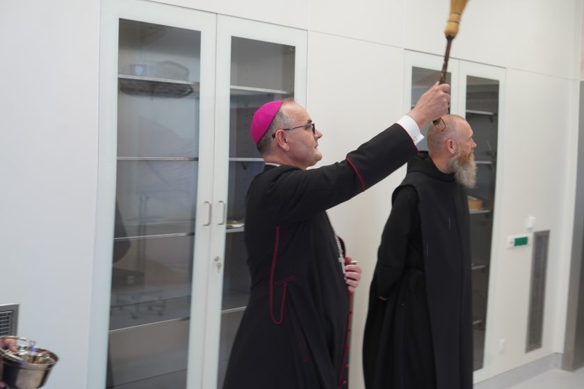 Zdjęcie przedstawia biskupa w białych szatach liturgicznych z fioletową piuską, który unosi kropidło i błogosławi nowo otwarte pomieszczenia szpitalne. Obok niego stoi zakonnik Zakonu Bonifratrów w czarnym habicie. W tle znajdują się wysokie szafy medyczne z przeszklonymi drzwiami oraz jasne ściany nowoczesnego wnętrza. W lewym dolnym rogu kadru widoczny jest fragment naczynia z wodą święconą. Zdjęcie dokumentuje moment poświęcenia nowych przestrzeni szpitalnych
