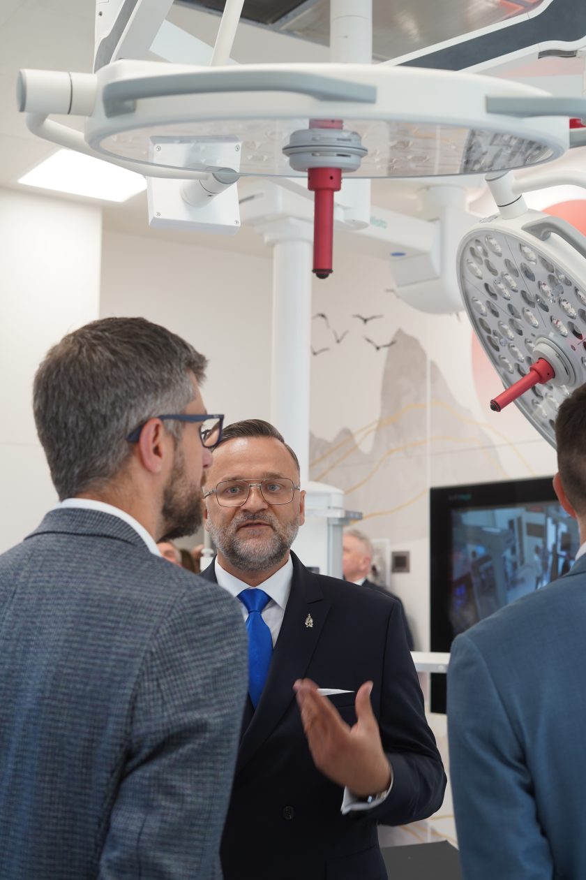 Zdjęcie przedstawia grupę uczestników uroczystości zwiedzających nowoczesną salę operacyjną. W centrum znajduje się mężczyzna w granatowym garniturze z niebieskim krawatem, który prowadzi rozmowę i gestykuluje podczas prezentacji wyposażenia. Nad uczestnikami widoczne są nowoczesne lampy operacyjne z panelami LED oraz systemy sufitowe do zawieszania sprzętu medycznego. W tle znajduje się monitor oraz dekoracyjna grafika przedstawiająca górski krajobraz z lecącymi ptakami. Całość ukazuje nowoczesny charakter zmodernizowanego bloku operacyjnego