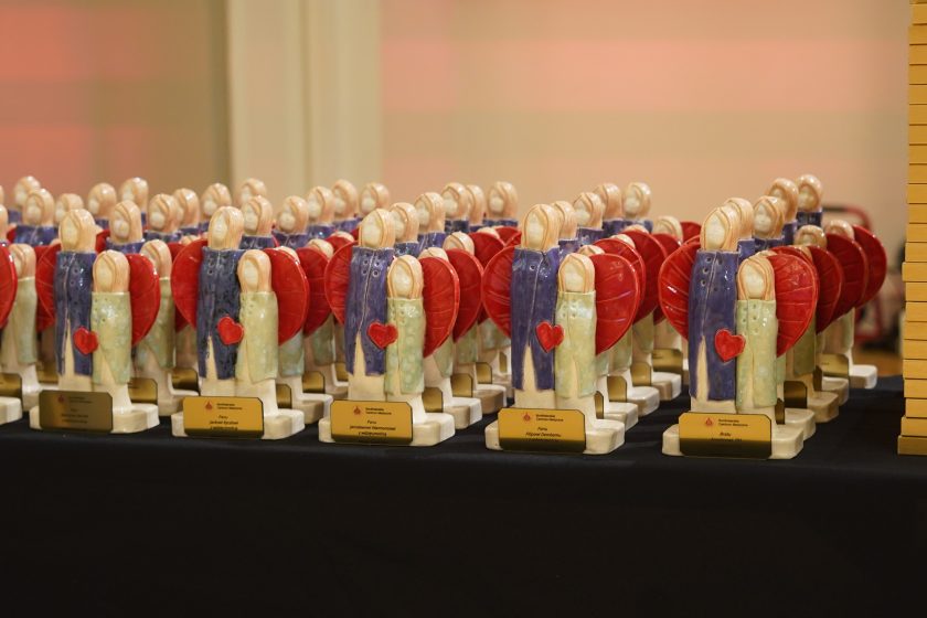 Zdjęcie przedstawia kilkadziesiąt ceramicznych statuetek ustawionych w równych rzędach na czarnym stole przed rozpoczęciem uroczystej gali. Każda statuetka przedstawia dwie stylizowane postacie obejmujące czerwone serce na tle dużych czerwonych skrzydeł. U podstawy znajdują się złote tabliczki z nazwiskami wyróżnionych osób oraz logo Bonifraterskiego Centrum Medycznego. W tle widoczne są kolejne rzędy identycznych nagród oraz ciepło oświetlona sala
