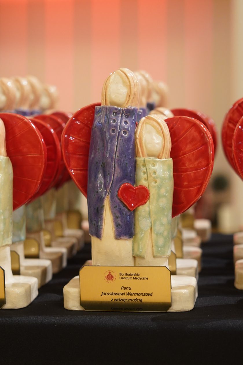 Zdjęcie przedstawia zbliżenie jednej z ceramicznych statuetek przygotowanych dla laureatów uroczystości. Figurka przedstawia dwie stylizowane postacie stojące obok siebie na tle czerwonych skrzydeł, z czerwonym sercem umieszczonym pomiędzy nimi. U podstawy znajduje się złota tabliczka z napisem: „Bonifraterskie Centrum Medyczne. Panu Jarosławowi Warmonsowi z wdzięcznością. Katowice, czerwiec 2026”. W tle widoczne są kolejne identyczne statuetki ustawione w rzędach